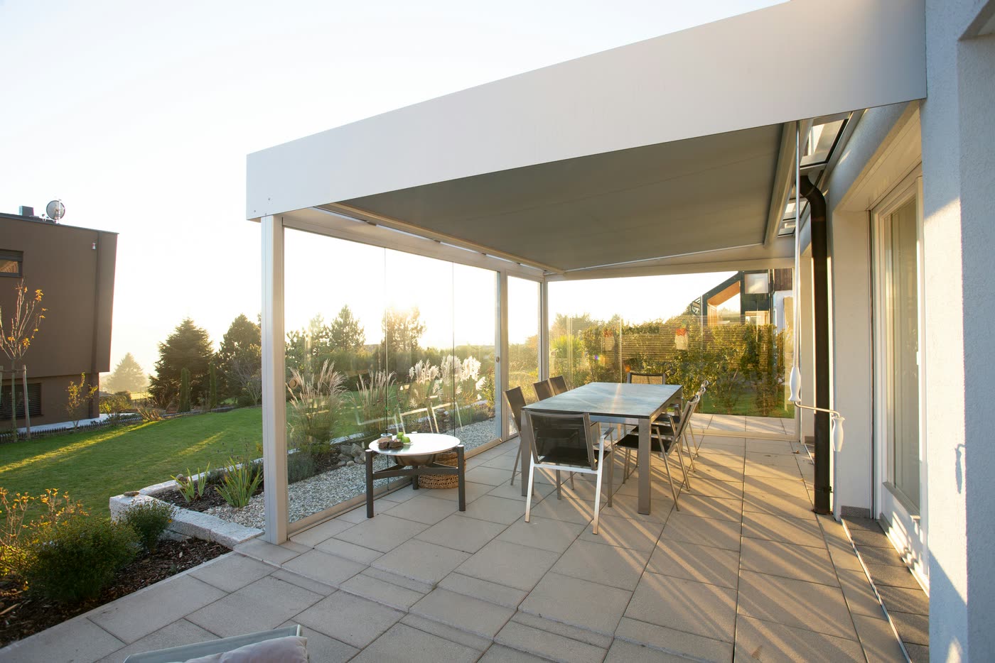 Covered terrace extension with a glass balustrade and outdoor dining setting looking out over a lawn in the evening light