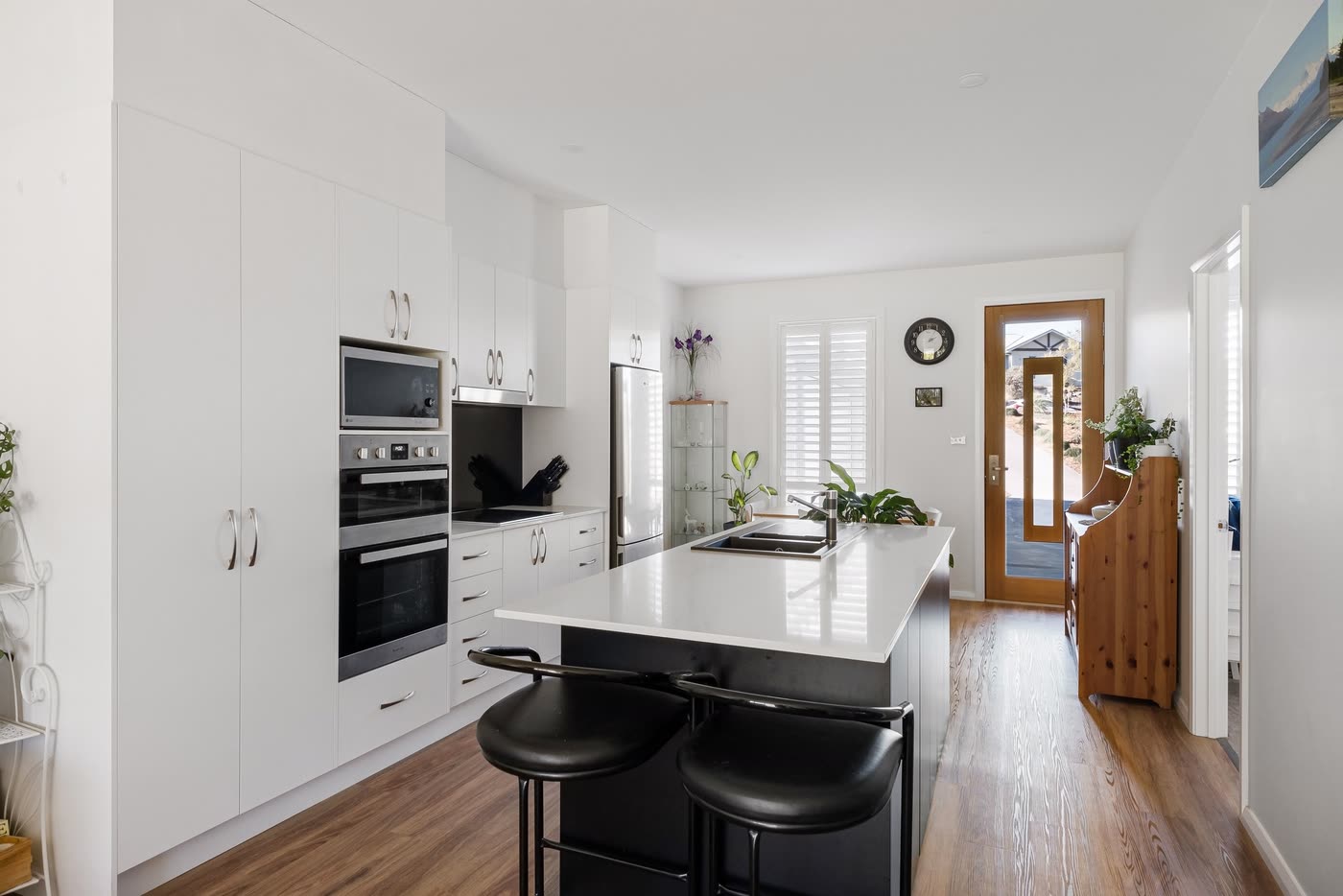 White galley kitchen renovation with a dark island bench, integrated wall ovens and timber floorboards